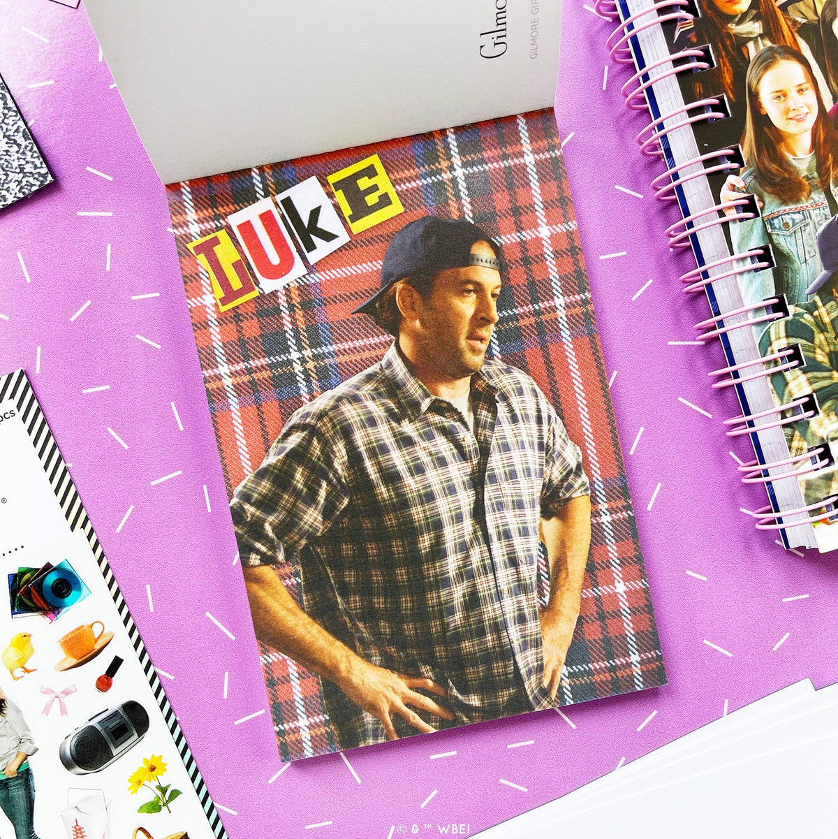 A styled flat-lay of a postcard in the postcard pack featuring a man in a plaid shirt and cap, placed on a purple surface with other stationery items around it. The card has a red, blue, and black plaid background with the word 'LUKE' in large colorful le