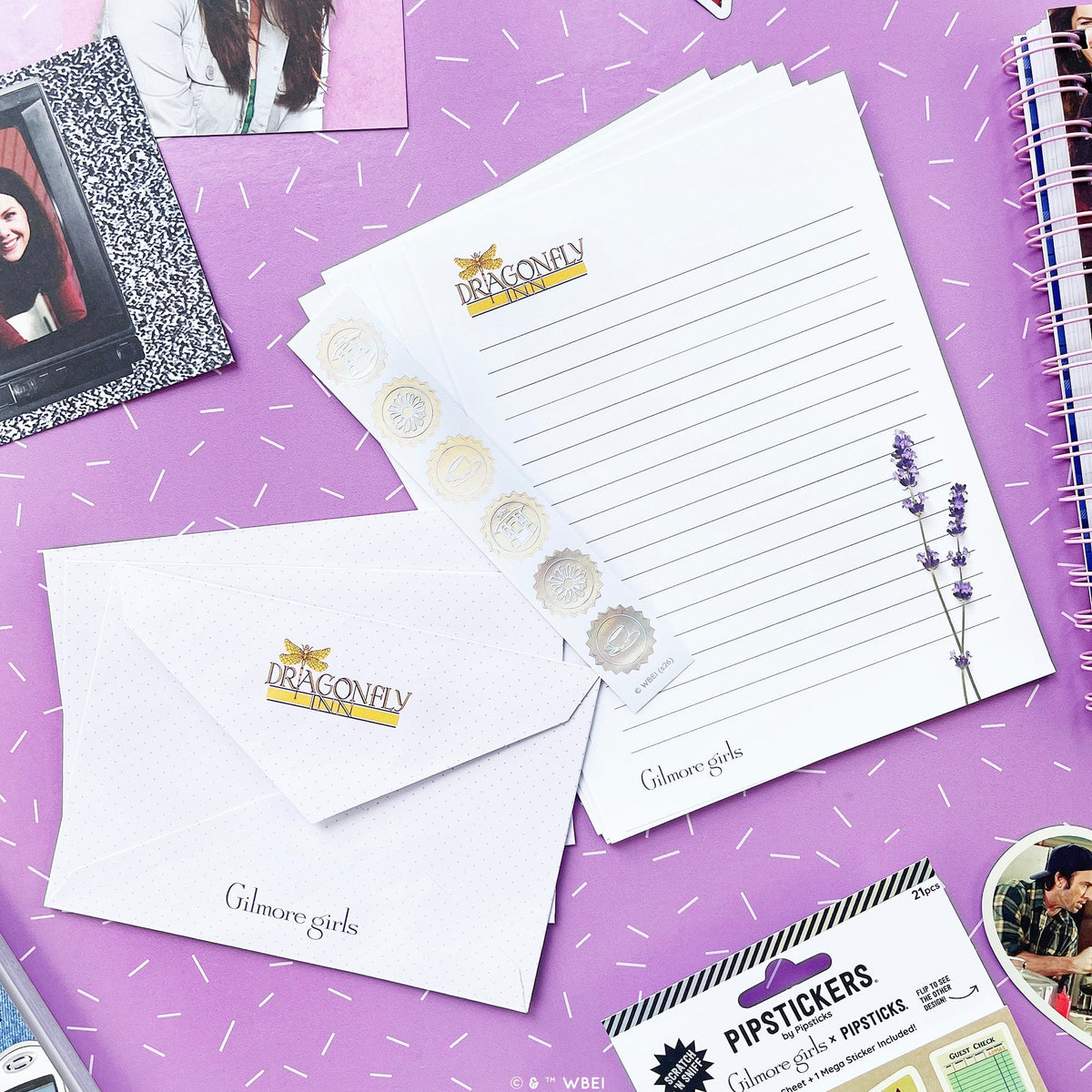 A styled flat-lay of Dragonfly Girls stationery on a purple background with white confetti. The set includes a lined letterhead pad, a matching envelope, a sticker strip with lavender illustrations, and a package of lip stickers.
